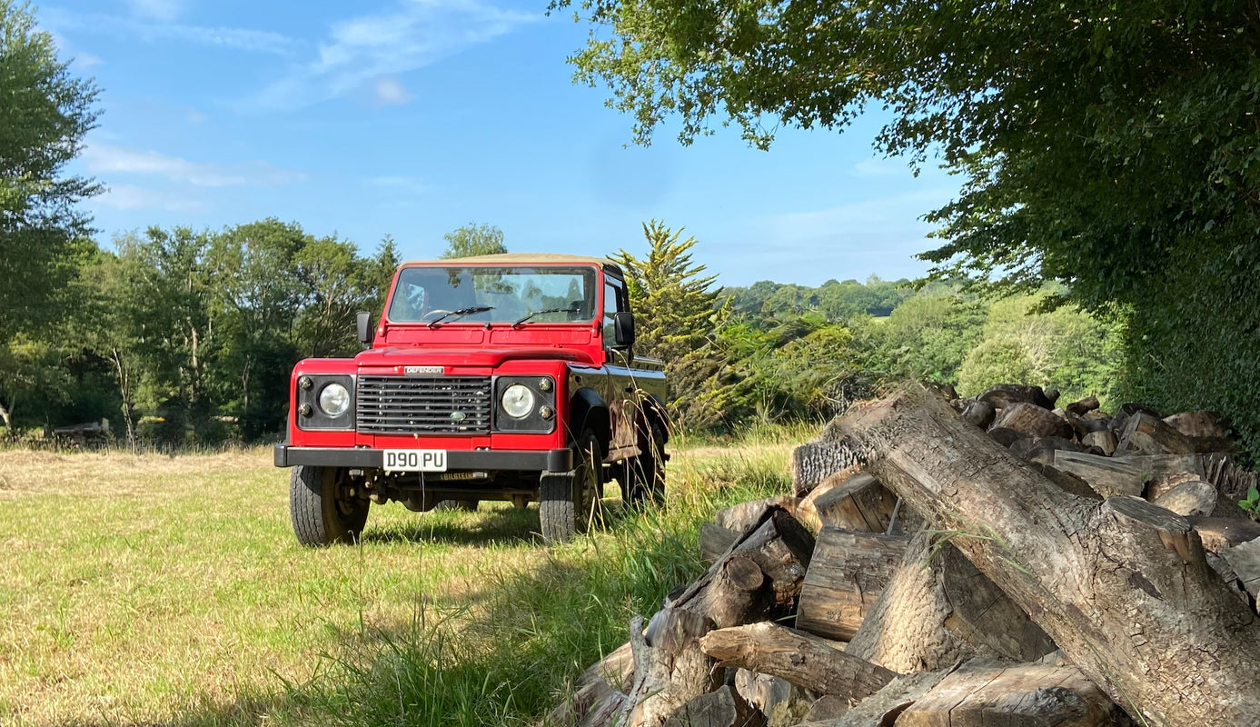 Our classic Land Rover Defender red pick up truck parked in a field next to a pile of logs